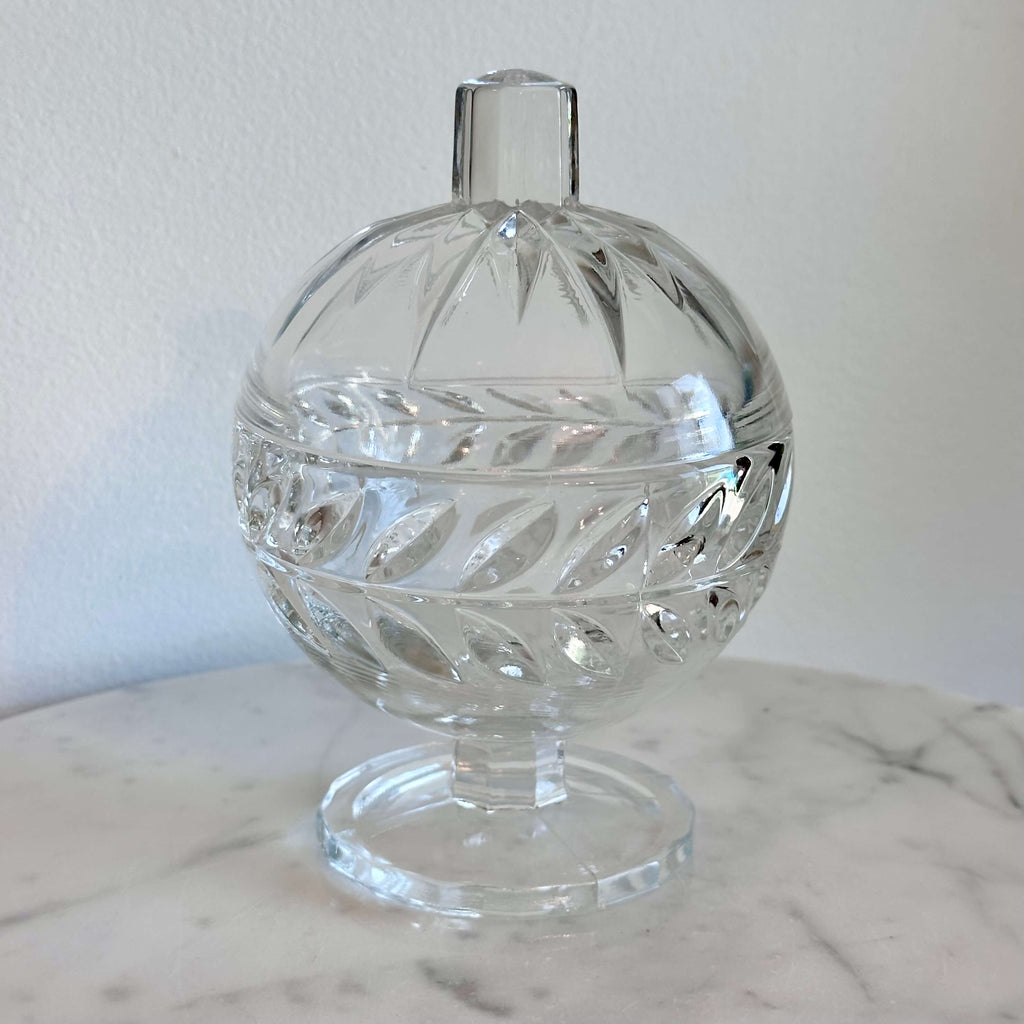 A clear cut-glass  pedestal container with a rounded body, decorative facets, and a small cylindrical top, shown on a marble surface against a plain light wall.