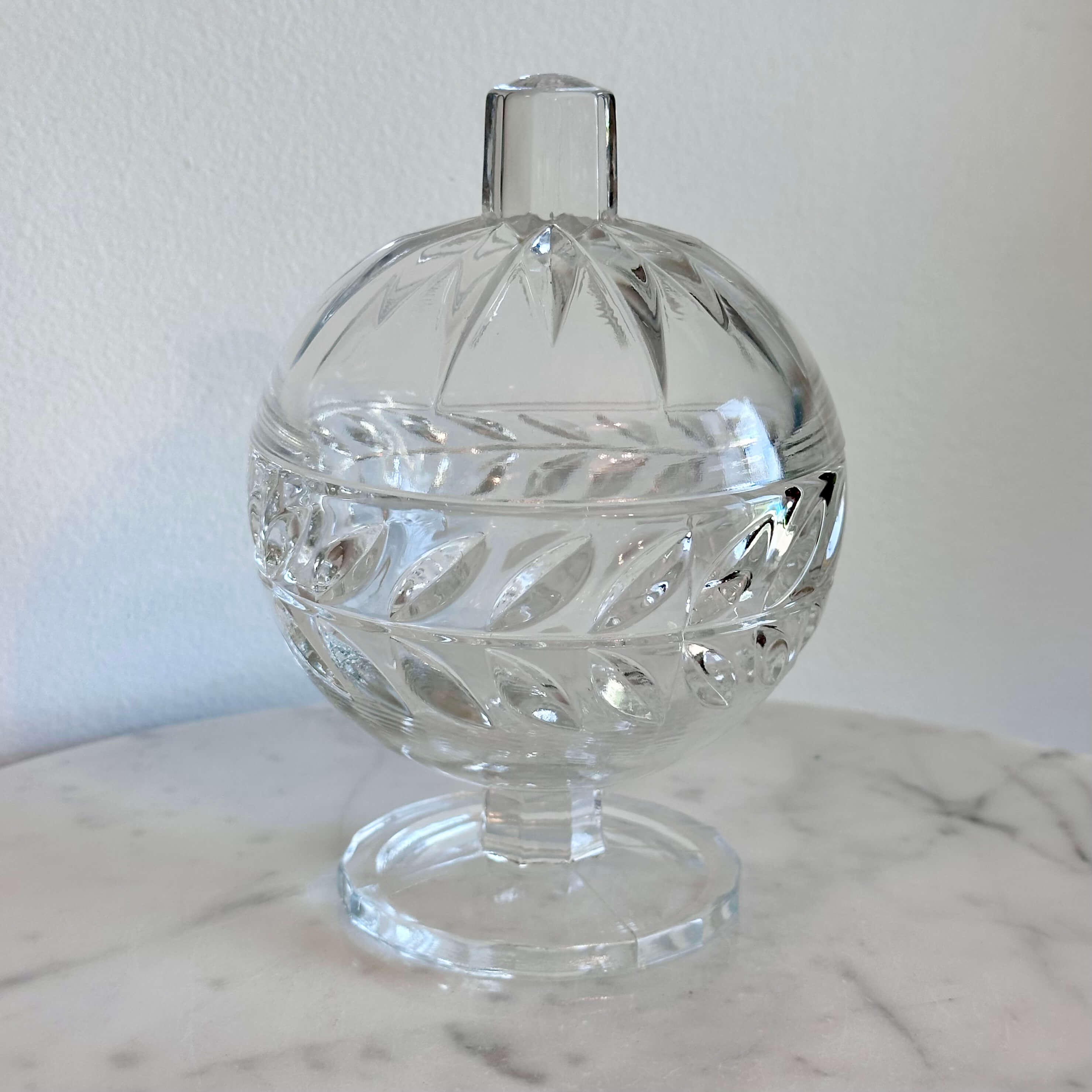 A clear cut-glass  pedestal container with a rounded body, decorative facets, and a small cylindrical top, shown on a marble surface against a plain light wall.