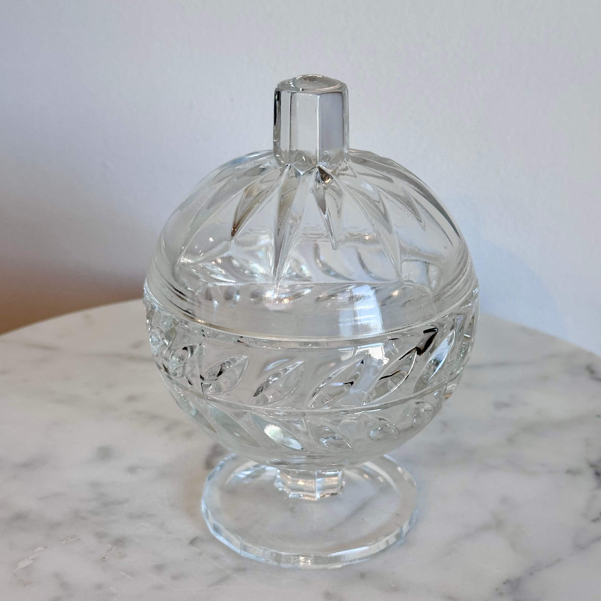 A clear cut-glass  pedestal container with a rounded body, decorative facets, and a small cylindrical top, shown on a marble surface against a plain light wall.