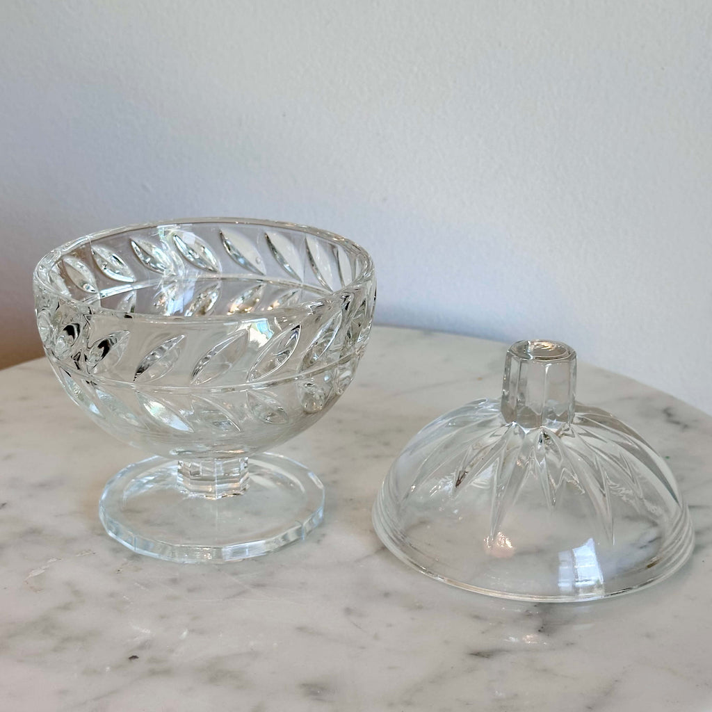 A clear cut-glass  pedestal container with a rounded body, decorative facets, and a small cylindrical top, shown on a marble surface against a plain light wall.