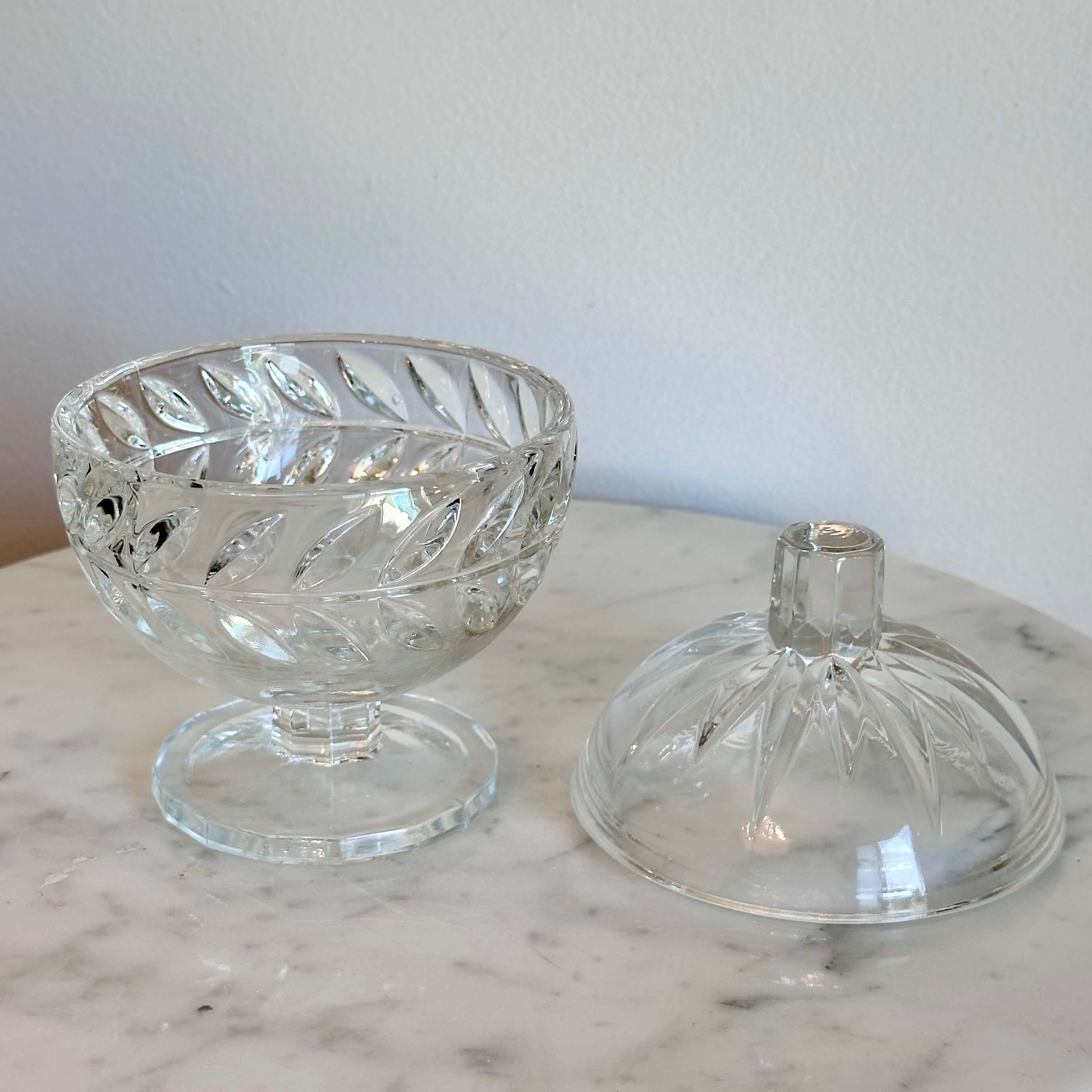 A clear cut-glass  pedestal container with a rounded body, decorative facets, and a small cylindrical top, shown on a marble surface against a plain light wall.