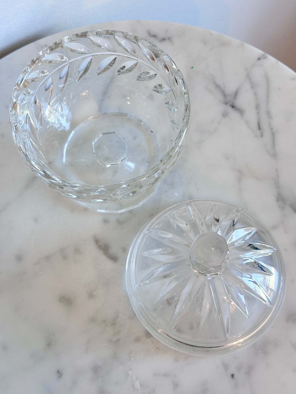 A clear cut-glass  pedestal container with a rounded body, decorative facets, and a small cylindrical top, shown on a marble surface against a plain light wall.